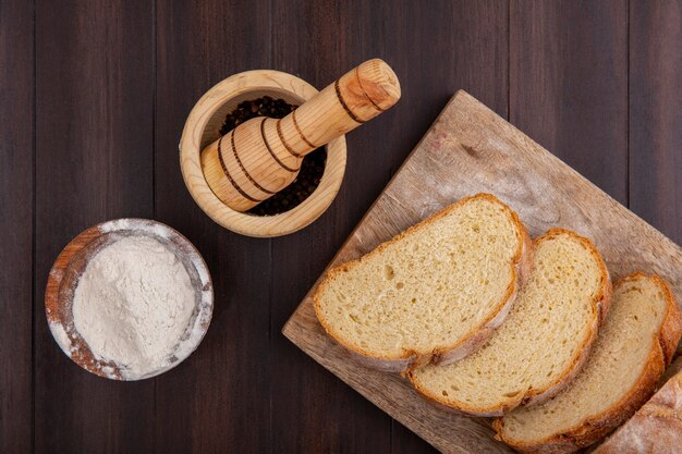 Top view of sliced crusty bread on cutting board and flour in bowl with black pepper in garlic crusher on wooden background