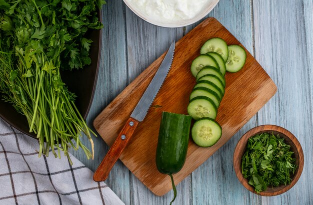 Top view of sliced cucumbers on a board with a knife and greens on a gray surface