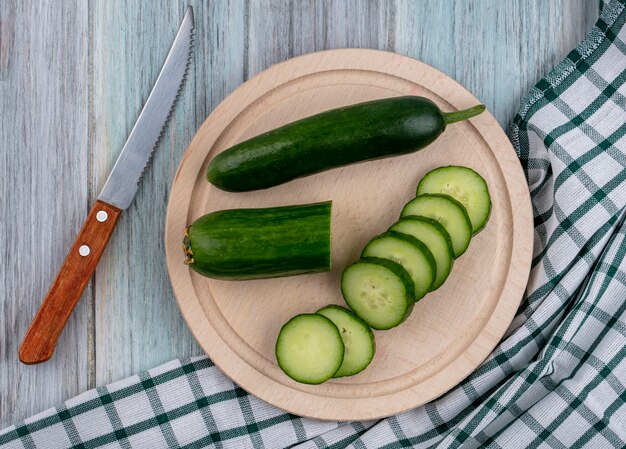 Top view of sliced cucumbers on a stand with a knife on a checkered towel on a gray surface