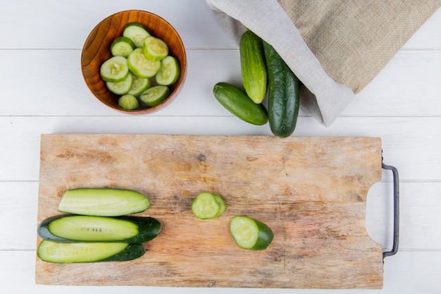 Top view of sliced and cut cucumber on cutting board with bowl of cucumber slices and cucumbers spilling out of sack on wooden surface