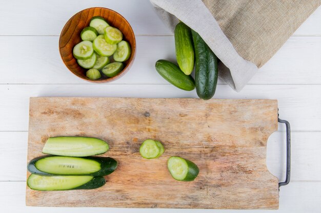 Top view of sliced and cut cucumber on cutting board with bowl of cucumber slices and cucumbers spilling out of sack on wooden surface