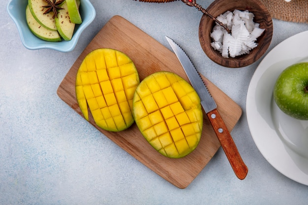 Top view of sliced fresh mango on a wooden kitchen board with knife and chopped apples in a white bowl and pulps of coconut in a wooden bowl on white surface