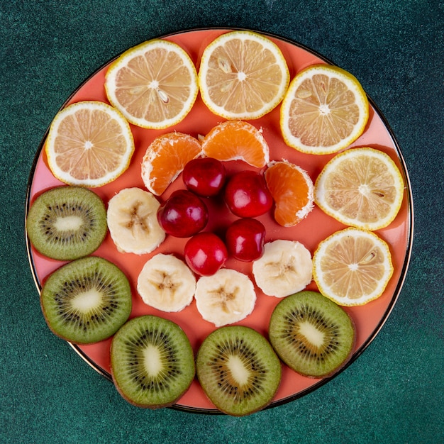 Вид сверху нарезанные фрукты киви лимон банан мандарин и красная вишня на тарелку на темном