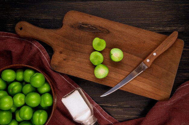 Top view of sliced green plums with a kitchen knife on a wooden cutting board with a bowl filled with green plums on dark rustic table