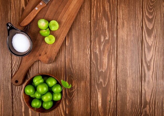 Top view of sliced green plums with salt and kitchen knife on a wooden cutting board and plums in a bowl on rustic wooden table with copy space