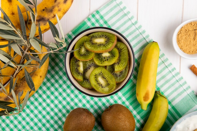 Top view of sliced kiwi in a bowl, fresh banana fruits on white wood
