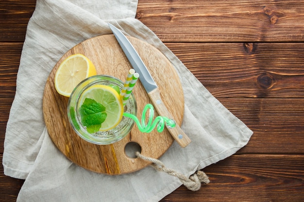 Top view sliced lemon in bowl with cutting board, straw, white cloth, wooden knife on wooden surface. vertical