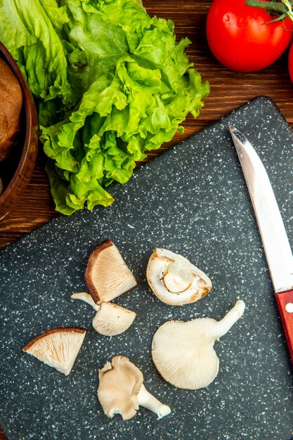 Top view of sliced mushrooms with lettuce tomato and kitchen knife on black board on wood