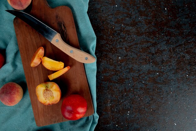 Top view of sliced peach with knife on cutting board with whole peaches on cloth on brown and black surface with copy space