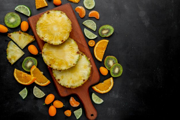 Top view of sliced pineapples on cutting board with other citrus fruits around on black surface