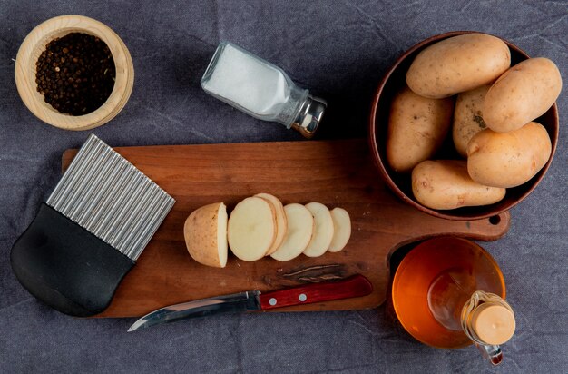 Top view of sliced potato and knife with potato chip cutter on cutting board with other ones in bowl salt black pepper butter on gray cloth