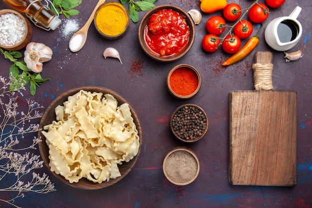 Top view sliced raw dough with red tomatoes and different seasonings on dark desk dough pasta meal food vegetable