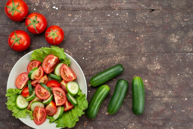 Top view sliced tomatoes with cucumbers inside white plate with green salad along with fresh vegetables on brown, food vegetable fresh salad