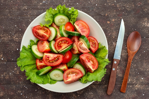 Pomodori affettati vista superiore con i cetrioli dentro il piatto bianco con insalata verde su marrone, sald fresco di verdure del pranzo dell'alimento