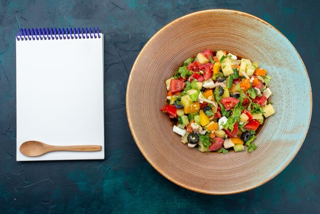 Top view sliced vegetable salad peppered inside plate with notepad on the dark-blue desk