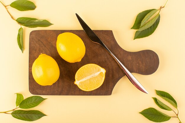 Вид сверху нарезанный цельный лимон свежей сочной спелости вместе с серебряным ножом и зелеными листьями на коричневом деревянном столе и кремовом фоне цитрусового апельсина