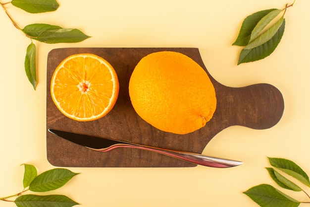 A top view sliced whole orange fresh juicy mellow along with silver knife and green leaves on the brown wooden desk and cream background citrus orange