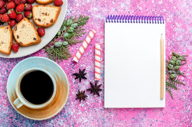 Free photo top view sliced yummy cakes with fresh red strawberries notepad and cup of coffee on light-pink desk