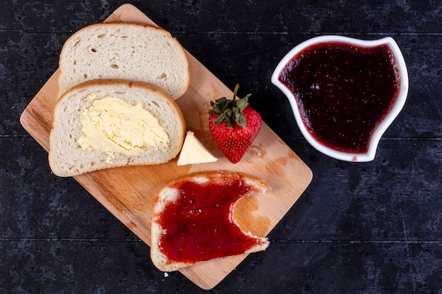 Vista dall'alto fette di pane e burro con una fetta di pane con marmellata a bordo con fragole e marmellata in un piattino