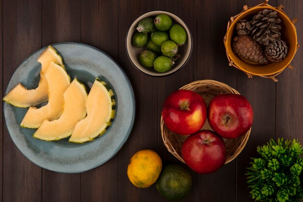 Вид сверху ломтиков дыни на тарелке с фейхоа на миске с яблоками на ведре с мандаринами, изолированными на деревянной стене