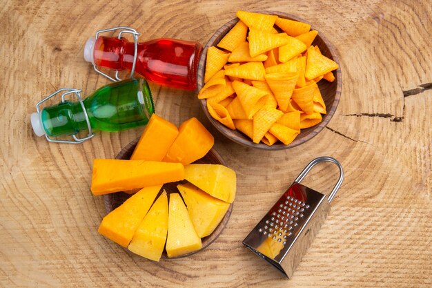 Top view slices of cheese and chips in bowls grater green and red small bottles on wooden ground