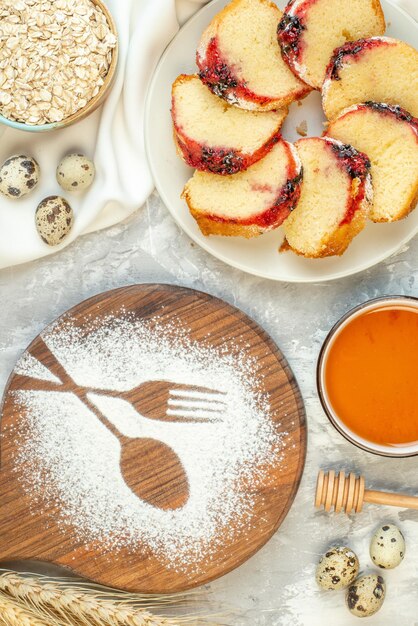 Вид сверху ломтики торта с джемом на тарелке, мед и овес в мисках, вилка и ложка перепелиных яиц, отпечатанные на деревянной доске с мукой на столе
