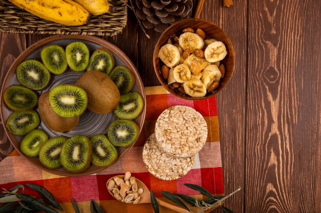 Вид сверху кусочки киви на тарелку и нарезанные бананы с миндалем в деревянной миске, деревянной ложкой с арахисом и рисовые крекеры на деревенском с копией пространства