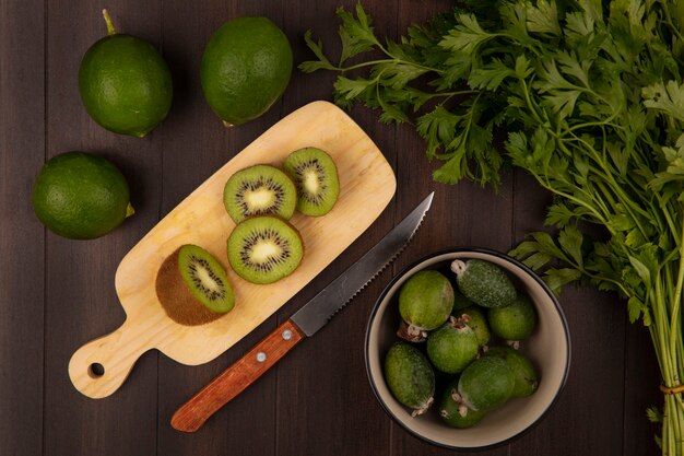 Top view of slices of kiwi on a wooden kitchen board with knife with feijoas on a bowl with parsley and limes isolated on a wooden wall