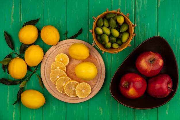 Top view of slices of lemons on a wooden kitchen board with red apples on a bowl with kinkans on a bucket with lemons isolated on a green wooden wall