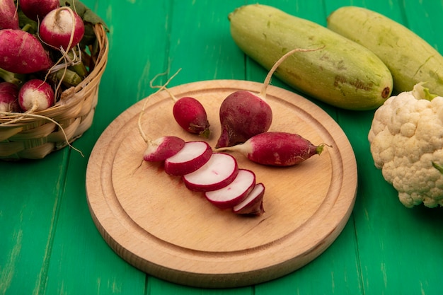 Top view of slices of radish isolated on a wooden kitchen board with whole radishes on a bucket with zucchinis and cauliflower isolated on a green wooden wall