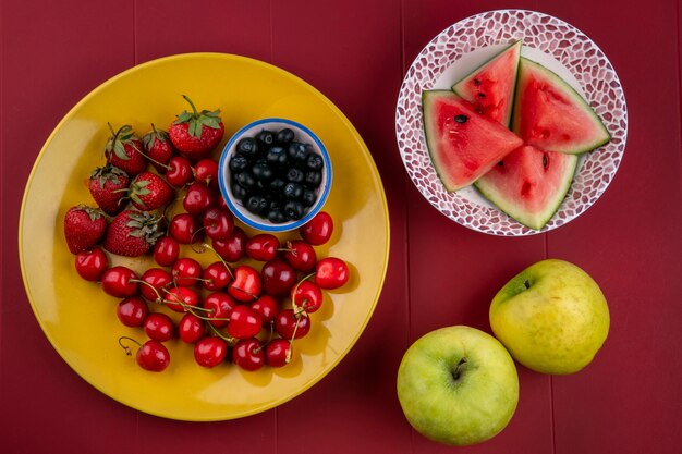 Вид сверху ломтики арбуза с черникой, клубникой, вишней на тарелке и яблоками на красном фоне