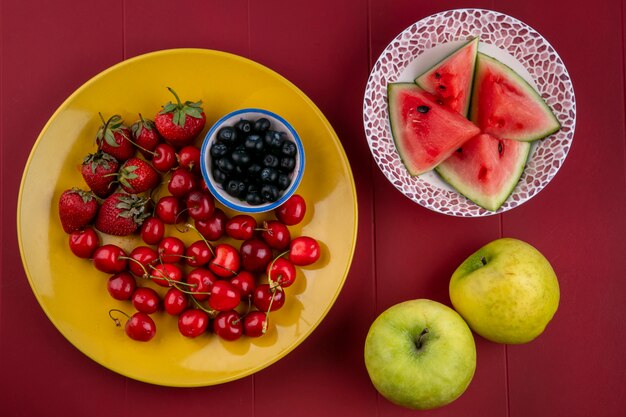 Top view slices of watermelon with blueberries strawberries cherries on a plate and apples on a red background