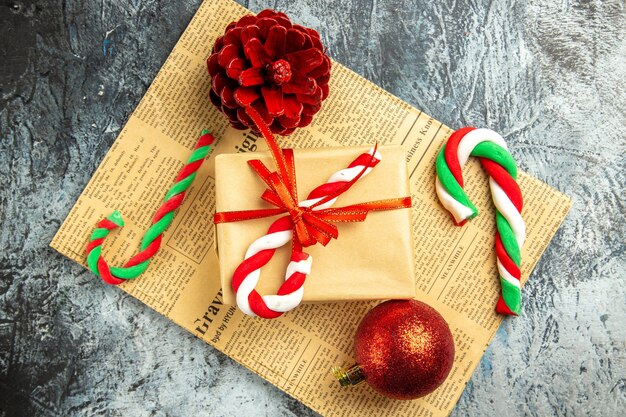 Вид сверху небольшой подарок, перевязанный красной лентой, рождественские конфеты на газете на серой поверхности