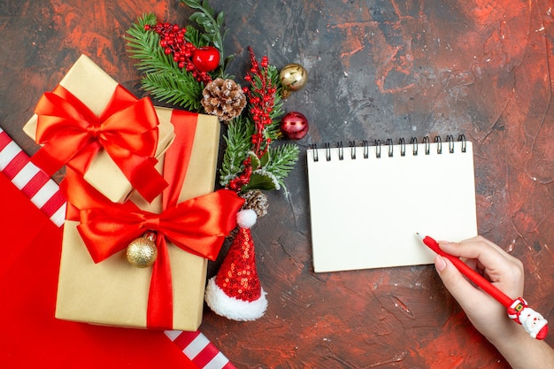 Top view small gifts tied with red ribbon santa hat xmas tree branch notepad red pen on dark red table