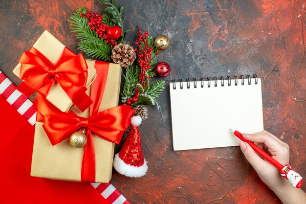 빨간색 리본 산타 모자 크리스마스 트리 분기 메모장 빨간색 펜으로 묶인 상위 뷰 작은 선물 어두운 빨간색 테이블에