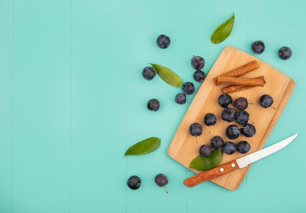 Top view of the small sour blackish fruit sloes on a wooden kitchen board with cinnamon sticks with knife on a blue background with copy space