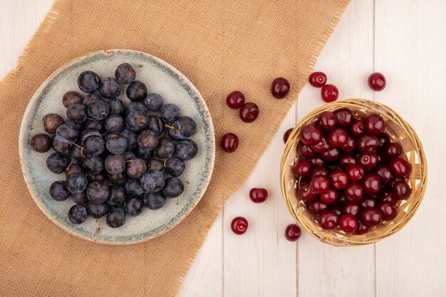 Top view of the small sour blue-black fruit sloes on a bowl on a sack cloth with red cherries on a bucket on a white background