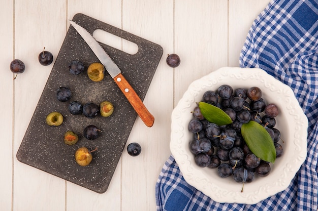 Foto gratuita vista dall'alto delle piccole prugnole di frutta blu-nere acide su una ciotola con fette di prugnole su un tagliere da cucina con coltello su sfondo bianco