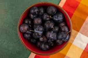 Foto gratuita vista dall'alto delle piccole prugnole di frutta blu-nere acide su una ciotola rossa su una tovaglia a quadretti su uno sfondo verde