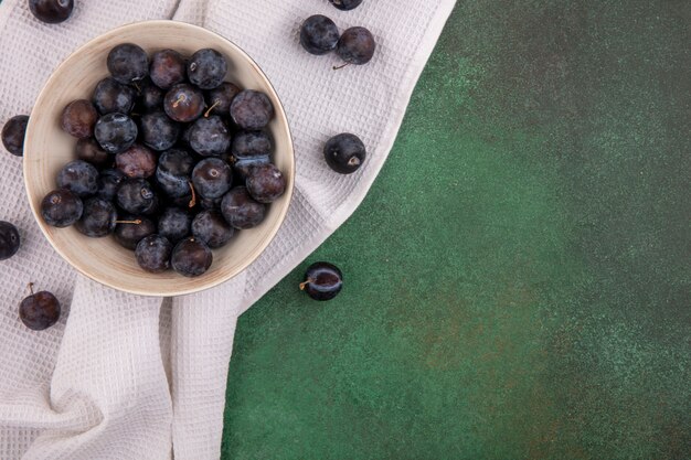 Top view of the small sour blue-black fruit sloes on a white bowl on a tablecloth on a green background with copy space