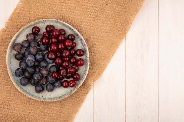 Top view of the small sour blue-black fruit sloes with red cherries on a plate on a sack cloth on a white background with copy space