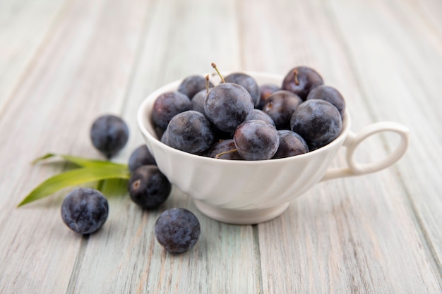 Top view of the small sour blue-black sloe on a white cup with leaves on a grey wooden background