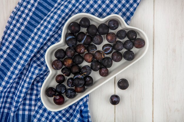 Top view of the small sour blue-black sloes on a white bowl on a blue checked tablecloth on a grey wooden background