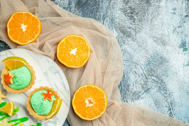 Top view small tarts with green pastry cream and lemon slice on plate on beige shawl cut oranges on dark table free space