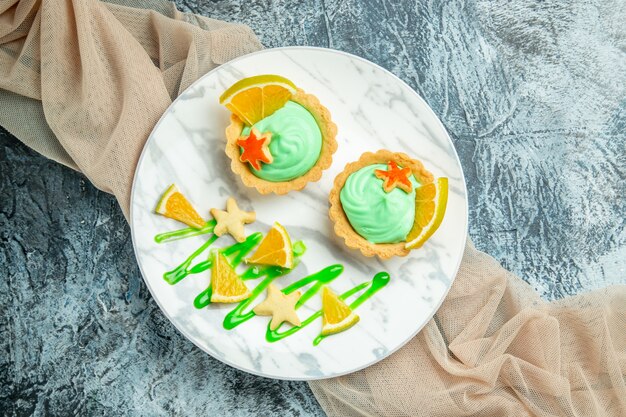 Top view small tarts with green pastry cream and lemon slice on plate on beige shawl on dark table