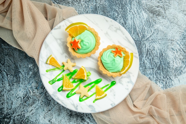 Top view small tarts with green pastry cream and lemon slice on plate on beige shawl on dark table