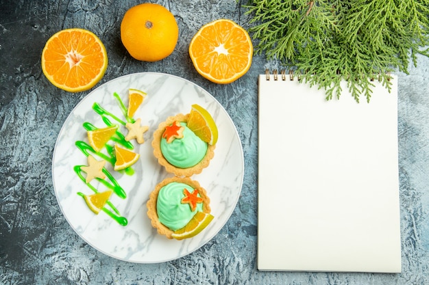 Vista dall'alto piccole crostate con crema pasticcera verde e fetta di limone su taccuino di arance tagliate piatto sul tavolo scuro