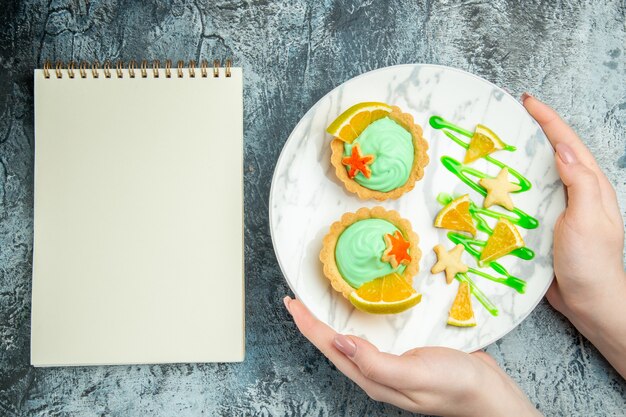Top view small tarts with green pastry cream and lemon slice on plate in woman hands notebook on grey table