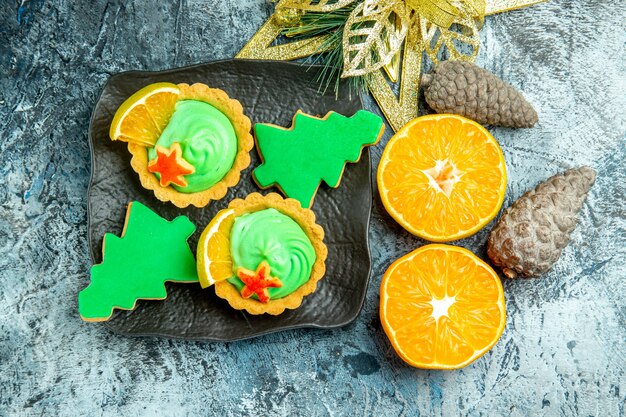 Top view small tarts with green pastry cream xmas tree biscuits on black plate xmas ornament cut oranges on grey table