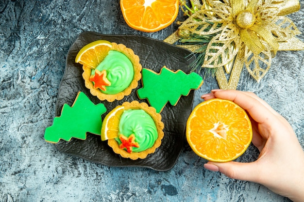 Top view small tarts xmas tree biscuits on black plate xmas ornament cut orange in female hand on grey table